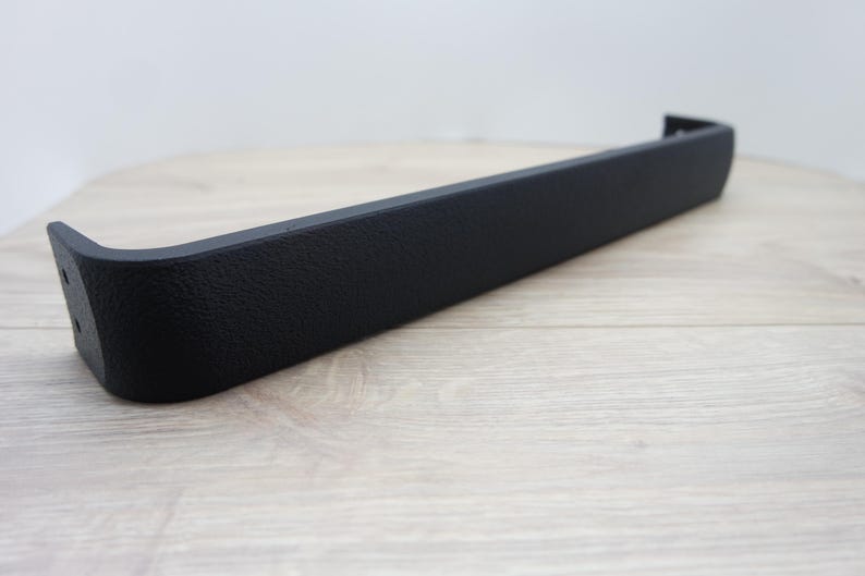 May include: A black, textured, rectangular automotive part with a curved end and two holes. The part is set against a light wood-grain surface and a plain white background. The part appears to be made of a durable material.