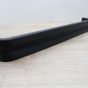 May include: A black, textured, rectangular shelf bracket with a curved corner. The bracket is made of a solid material and is designed to support a shelf. The bracket is shown on a light wood surface.