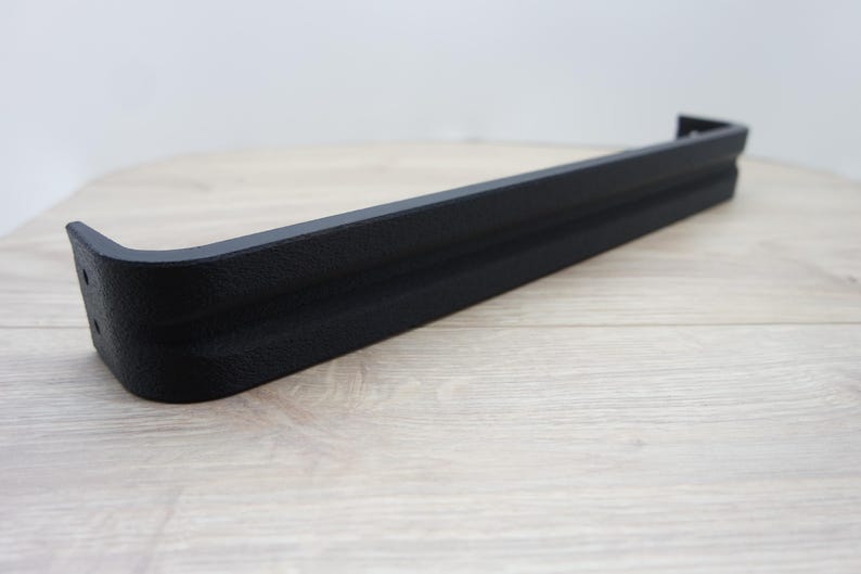 May include: A black, textured, rectangular shelf bracket with a curved corner. The bracket is made of a solid material and is designed to support a shelf. The bracket is shown on a light wood surface.