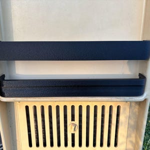 May include: A section of a vintage camper interior, featuring a cream-colored panel with two black textured trim pieces. Below, a gold-colored vent with vertical slats and a central lock is visible.