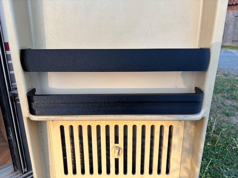 May include: A section of a vintage camper interior, featuring a cream-colored panel with two black textured trim pieces. Below, a gold-colored vent with vertical slats and a central lock is visible.