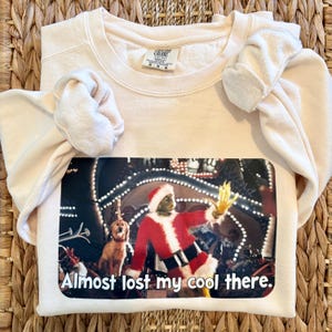 Puede incluir: Sudadera de color crema con un gráfico de un personaje con un traje de Papá Noel y el texto "Almost lost my cool there." El gráfico es una escena de una película navideña. La sudadera tiene mangas largas y cuello redondo.