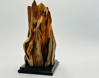 May include: A wooden sculpture of a tree trunk with a natural wood finish. The sculpture is on a black base.