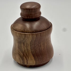 May include: A handcrafted wooden container with a lid. The container is a rich brown color and has a rounded shape with a slightly flared base. The lid is a smaller, rounded shape that fits snugly on top. The wood grain is visible.