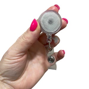 May include: A clear retractable badge holder with a silver clip and a white glitter-filled circle. The holder is attached to a clear plastic strap.