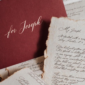 May include: A burgundy envelope with the words "for Joseph" in elegant white script, resting on a collection of aged, handwritten letters. The paper has torn edges, creating a vintage aesthetic.