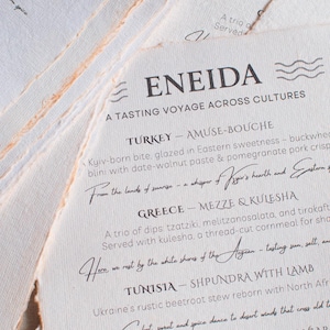 May include: A stack of textured paper menus with a torn edge, featuring elegant black script. The menus are for a tasting voyage across cultures, with dishes from Turkey, Greece, Tunisia, and Sicily. The word "ENEIDA" is at the top.