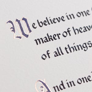 May include: Close-up of a page with elegant calligraphy. The text, in a classic font, reads "We believe in one...maker of heaven...of all things." The initial letters are in dark blue with gold accents, set against a light background.