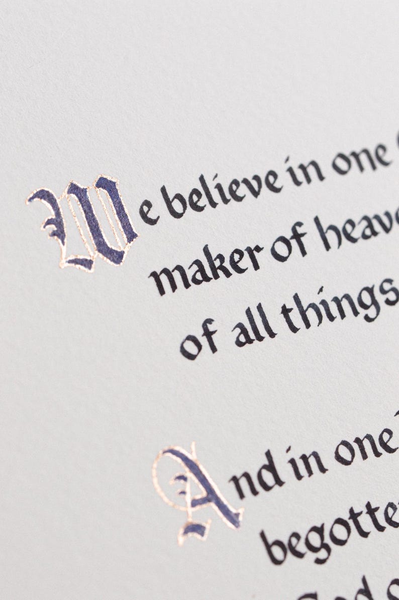 May include: Close-up of a page with elegant calligraphy. The text, in a classic font, reads "We believe in one...maker of heaven...of all things." The initial letters are in dark blue with gold accents, set against a light background.