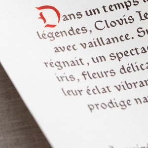 May include: Close-up of a white paper with French text in a decorative font. The first letter is a large, ornate red 'D'. The text is in brown ink, with words like 'l&eacute;gendes' and 'Clovis' visible.