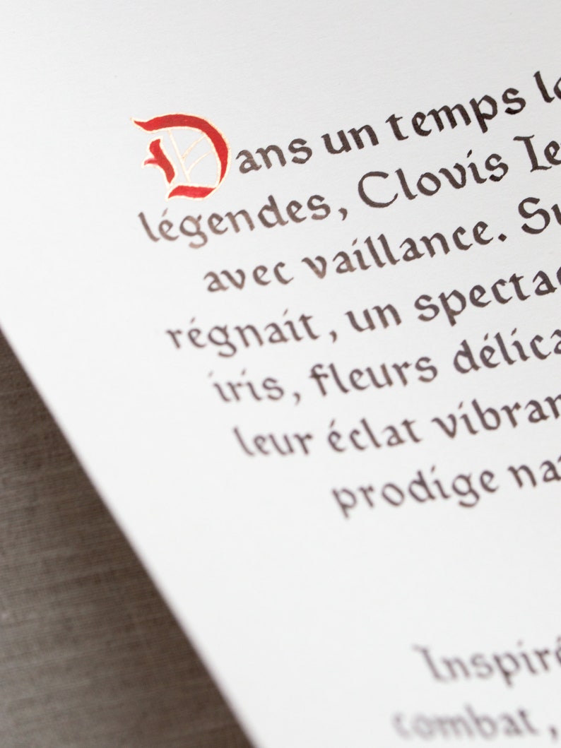 May include: Close-up of a white paper with French text in a decorative font. The first letter is a large, ornate red 'D'. The text is in brown ink, with words like 'l&eacute;gendes' and 'Clovis' visible.