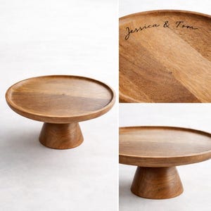 Peut inclure: Un présentoir à gâteau rond en bois avec un socle. Le présentoir est en bois brun clair et a une surface lisse et polie. Le dessus du présentoir porte l'inscription "Jessica & Tom" en écriture cursive.