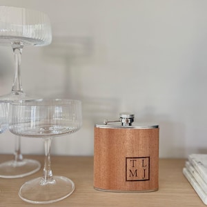 Könnte beinhalten: Eine rechteckige Holzflasche mit silbernem Verschluss und Scharnier. Auf der Vorderseite sind die Initialen "TL MJ" in einem Quadrat eingraviert. Im Hintergrund sind zwei Champagnergläser zu sehen.