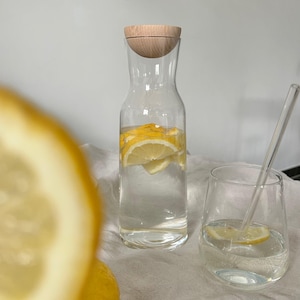 Könnte beinhalten: Eine klare Wasserkaraffe aus Glas mit Holzdeckel, gefüllt mit Wasser und Zitronenscheiben. Daneben steht ein passendes Glas mit einer Zitronenscheibe und einem durchsichtigen Strohhalm. Die Karaffe und das Glas stehen auf einer weißen Oberfläche.