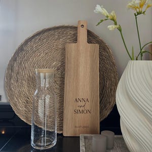 May include: A wooden cutting board with a handle, engraved with the names "ANNA und SIMON" and the date "31-07-2023". A clear glass carafe and a woven placemat are in the background. A white vase with flowers is on the right.