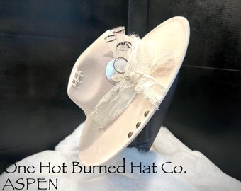 Pode incluir: Um chapéu de cowboy de feltro bege claro com uma guarnição de renda branca, um enfeite de metal prateado e um detalhe de pena marrom. O chapéu está em um suporte preto e o texto "One Hot Burned Hat Co. ASPEN" é visível abaixo do chapéu.