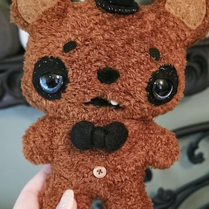 May include: A handmade brown plush bear with a black top hat and bow tie. The bear has large, expressive eyes, a button on its chest, and a single tooth. The plush toy is held in a person's hand.
