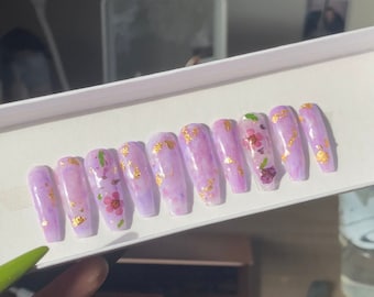 May include: Set of ten long, purple press-on nails with gold flakes and dried flowers. The nails are arranged in a row in a white box.