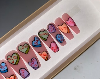 May include: A set of ten pink press-on nails with colorful heart designs. Each nail features a different Valentine's Day themed message, including "Love", "Miss U", "Kiss Me", "Hug Me", "Smile", "Cutie", "XO", "Be Mine", and "Bae".