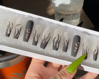 May include: A set of ten false nails with a frosted white base and black flame designs. Two of the nails have a small gold charm in the shape of a crescent moon.