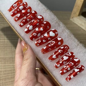 May include: Set of red and white press-on nails with a heart design. The nails are long and have a glossy finish.