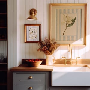 Puede incluir: Un fregadero de granja blanco con un grifo dorado y dos pomos. El fregadero está empotrado en una encimera de madera con gabinetes grises debajo. La encimera está iluminada por la luz del sol que entra por una ventana. Hay dos apliques de pared dorados a cada lado del fregadero y una impresión botánica enmarcada encima del fregadero.