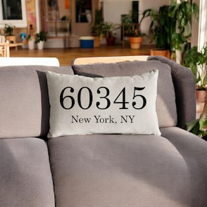 Könnte beinhalten: Ein rechteckiges Kissen mit dem Text "60345 New York, NY" in Schwarz auf hellem Beige gedruckt. Das Kissen liegt auf einem grauen Sofa.