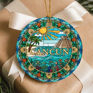 Może przedstawiać: Okrągła ozdoba świąteczna z kolorowym wzorem Cancun. Ozdoba przedstawia scenę na plaży z piramidą, palmami i napisem "CANCUN". Projekt jest obramowany obwódką w stylu witrażu ze złotym sznurkiem do zawieszania.