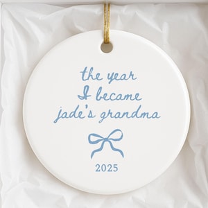 May include: A white ceramic ornament with the text "the year I became jade's grandma" in light blue script, a blue bow, and the year 2025. The ornament is in a white gift box with tissue paper and a gold string.
