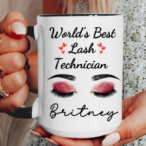 May include: White ceramic mug with a black rim and handle. The mug has a design of a pair of pink glitter eyelashes with the text "World's Best Lash Technician" and the name "Britney" below.