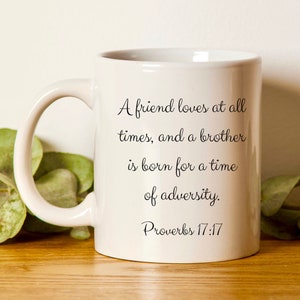 May include: White ceramic coffee mug with black text. The text is a bible verse from Proverbs 17:17 that reads: 'A friend loves at all times, and a brother is born for a time of adversity.'  The mug is shown on a wooden surface with green leaves.