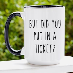 May include: White ceramic coffee mug with a black rim and handle. The mug has black text that reads "BUT DID YOU PUT IN A TICKET?"