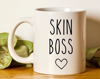 Könnte beinhalten: Eine weiße Keramiktasse mit den Worten "SKIN BOSS" in schwarzer Schrift und einem schwarzen Herzsymbol. Die Tasse hat einen gebogenen Henkel und ist von grünem Laub umgeben.