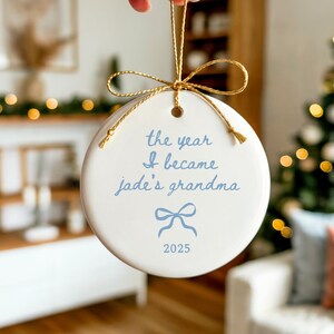 May include: A white ceramic Christmas ornament with gold string and bow. The ornament has the text "the year I became jade's grandma" in light blue, along with a bow graphic and the year 2025.