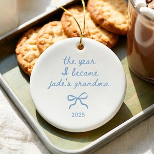 May include: A white ceramic ornament with the text "the year I became jade's grandma" in light blue script, a blue bow graphic, and the year 2025. The ornament hangs with a gold string. Cookies and a drink are in the background.