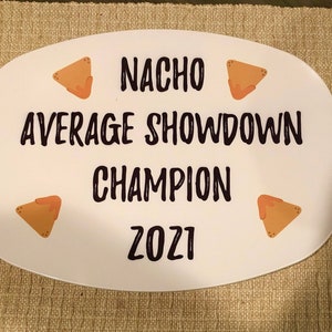 May include: An oval white serving platter featuring black text that reads NACHO AVERAGE SHOWDOWN CHAMPION 2021. The design includes four small illustrations of triangular tortilla chips with orange cheese sauce placed in the corners of the platter.
