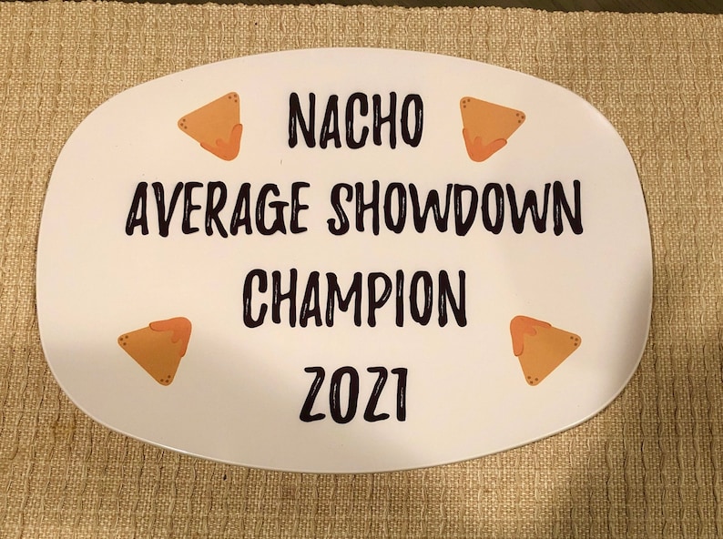 May include: An oval white serving platter featuring black text that reads NACHO AVERAGE SHOWDOWN CHAMPION 2021. The design includes four small illustrations of triangular tortilla chips with orange cheese sauce placed in the corners of the platter.