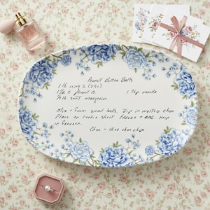 May include: An oval serving platter with a floral design in blue and white, featuring a handwritten recipe for peanut butter balls. The platter is surrounded by a floral pattern, with a perfume bottle and a ring box in the background.