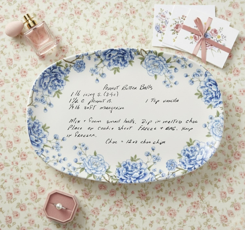 May include: An oval serving platter with a floral design in blue and white, featuring a handwritten recipe for peanut butter balls. The platter is surrounded by a floral pattern, with a perfume bottle and a ring box in the background.