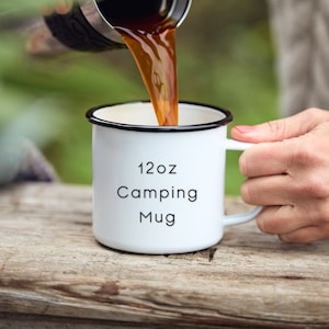 May include: White enamel camping mug with black rim and handle. The mug says "12oz Camping Mug". Coffee is being poured into the mug.