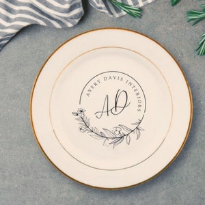May include: A white ceramic plate with a gold rim features a black and white logo with the text "Avery Davis Interiors" and the initials "AD" in a decorative font. The logo is surrounded by a wreath of stylized leaves and flowers.
