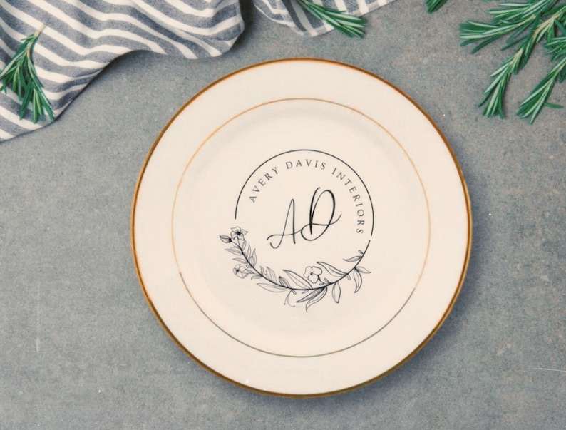 May include: A white ceramic plate with a gold rim features a black and white logo with the text "Avery Davis Interiors" and the initials "AD" in a decorative font. The logo is surrounded by a wreath of stylized leaves and flowers.