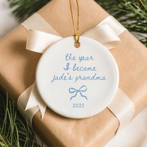 May include: A white ceramic ornament with the text "the year I became jade's grandma" in light blue script. A light blue bow graphic and the year 2025 are also featured. The ornament hangs from a gold string and is presented on a wrapped gift.
