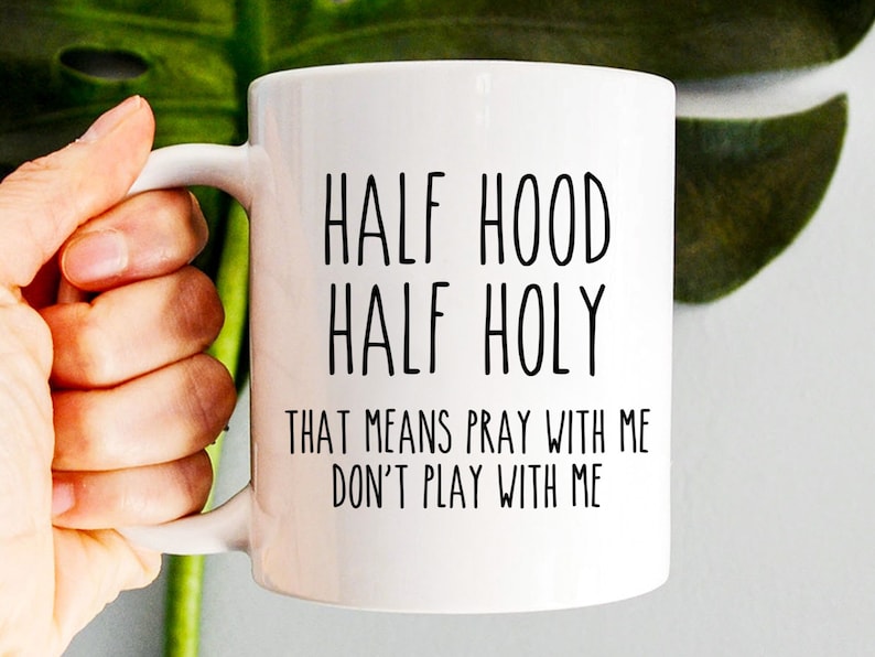 K&ouml;nnte beinhalten: Wei&szlig;e Keramiktasse mit schwarzem Text, der "Half Hood Half Holy That means pray with me don't play with me" lautet.