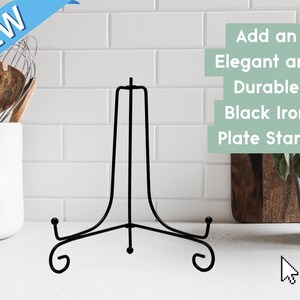 May include: A black iron plate stand with a central vertical support and two curved base arms featuring scroll-style feet. The text on the image reads: NEW, Add an Elegant and Durable Black Iron Plate Stand.