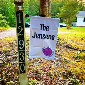 Op de afbeelding: Een witte tuin vlag met zwarte tekst die "The Jensens" luidt en een roze garenbal met een naald en de tekst "Yarn Addiction" in een cirkel. De vlag is bevestigd aan een houten paal met de nummers 12930.