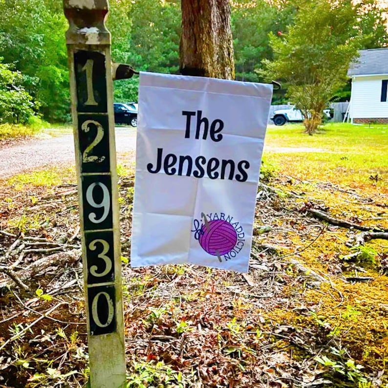 Op de afbeelding: Een witte tuin vlag met zwarte tekst die "The Jensens" luidt en een roze garenbal met een naald en de tekst "Yarn Addiction" in een cirkel. De vlag is bevestigd aan een houten paal met de nummers 12930.