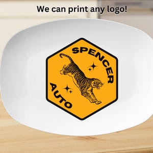May include: A white oval plate featuring a central yellow hexagon graphic with a black border. The graphic displays a leaping tiger, two four-pointed stars, and the black text "SPENCER AUTO". Above the plate, the text "We can print any logo!" appears.