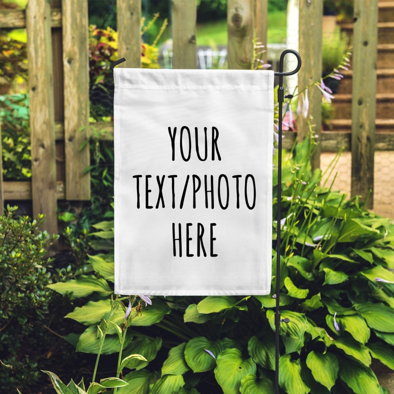 Op de afbeelding: Witte tuin vlag met zwarte tekst die "YOUR TEXT/PHOTO HERE" luidt. De vlag hangt aan een zwarte metalen paal in een tuin.