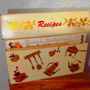 May include: Vintage recipe box with a cream-colored exterior and decorative kitchen-themed illustrations. The box is open, revealing recipe cards. The word "Recipes" is written in red on the lid. The box is made of metal.